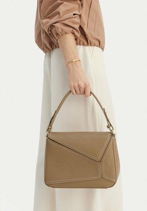 Mujer sostiene un bolso estructurado de cuero taupe con asa superior, con costuras blancas y herrajes dorados, vestida en tonos beige y crema.