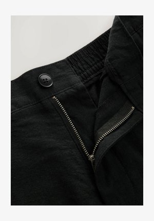 Juodos kelnes su užtrauktuku ir sagos uždarymu. Audinys atrodo tekstūruotas. Elastingas juosmuo matomas viršuje, su kontrasto užtrauktuku.