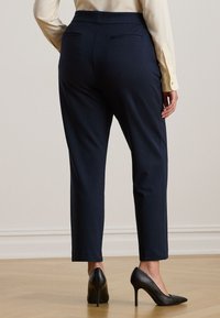 Námořnické kalhoty s tapered strihem vyrobené z hladké tkaniny, se středně vysokým pasem a dvěma zadními kapsami, v kombinaci s černými lodičkami na světlé dřevěné podlaze.