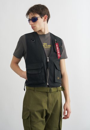 Νέος άνδρας που φοράει σκούρα γυαλιά ηλίου, μαύρο γιλέκο εργασίας με ταμπέλα "Αφαιρέστε Πριν την Πτήση", γκρι μπλουζάκι και χακί παντελόνι cargo σε λευκό φόντο.