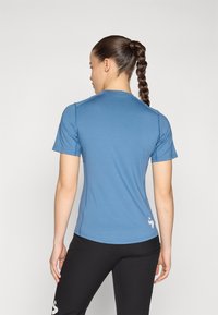 Modra športna majica s kratkimi rokavi, gladka tekstura, okrogla spodnja stran in majhna logotipna podrobnost na hrbtni strani. Vidne črne hlače.