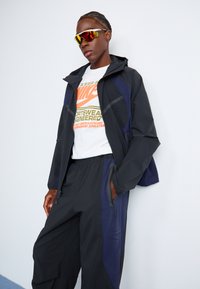 Black and navy windbreaker jacket with a hood, paired with black sweatpants featuring side zippers, and a white graphic T-shirt underneath.