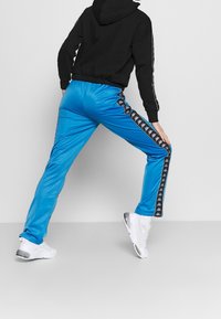 Blå idrottsbyxor med en glänsande yta, med svarta sidostråkar som har ett logomönster. I kombination med en svart huvtröja och vita sneakers.