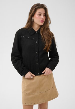 Giovane donna con capelli lunghi che indossa una giacca nera con borchie e una gonna beige con borchie su uno sfondo grigio uniforme.