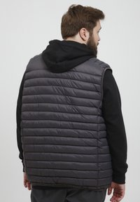 Vadderad väst i mörkgrått med horisontellt quiltat mönster, ärmlös design och en svart huva-tröja under.