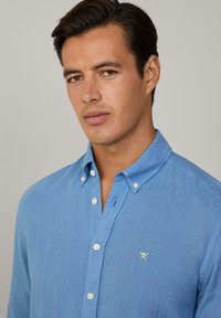 Camicia button-down azzurro chiaro realizzata in lino, con un piccolo logo giallo ricamato sul petto e un design con colletto classico.