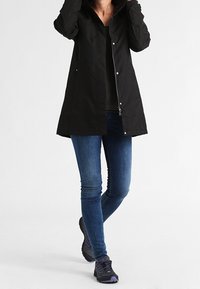 Osoba v černém dlouhém kabátu, modrých úzkých džínách a černých a fialových sportovních botách, s rukama blízko límce kabátu.