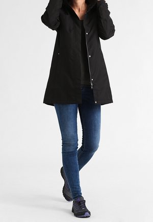 Osoba w czarnym długim płaszczu, niebieskich obcisłych dżinsach oraz czarnych i fioletowych butach sportowych, z rękami blisko kołnierza płaszcza.