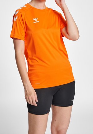 Person trägt ein orangefarbenes kurzärmeliges Hummel-Sportshirt und schwarze, eng anliegende Sportshorts vor einfarbigem Hintergrund.