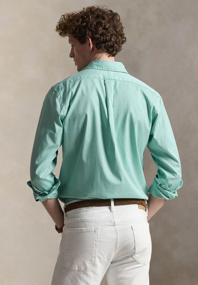 Άνδρας με πλάτη φοράει πουκάμισο μακρυμάνικο σε μέντα με γυρισμένα μανίκια, λευκό παντελόνι και καφέ ζώνη, μπροστά σε ουδέτερο φόντο.