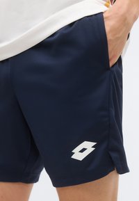 Άτομο που φοράει αθλητικό σορτς σε ναυτικό μπλε χρώμα με λευκό λογότυπο στο αριστερό πόδι, το χέρι τοποθετημένο στην δεξιά τσέπη.