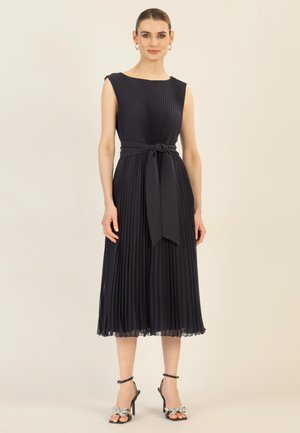 Marineblaues Plissee-Kleid mit engem Oberteil und Taillengürtel, das einen knielangen Rock und einen transparenten Saum hat. Getragen mit verzierten High-Heels.