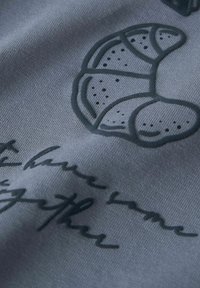 Grijze stof met een donker reliëfontwerp van een croissant en handgeschreven tekst. Gladde textuur met een subtiele glans.