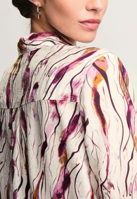 Vit blus med abstrakt lila, rosa och guldtryck. Har en krage och en flödande design som visar upp ett texturerat, lättviktsmaterial.