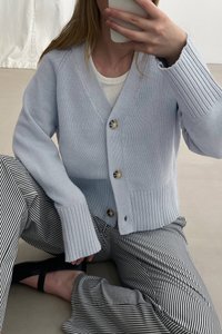 Cardigan lavorato a maglia azzurro chiaro con scollo a V, polsini a coste e bottoni frontali. Indossato sopra una maglietta bianca e abbinato a pantaloni a righe bianche e nere.