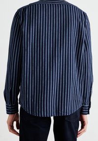 Bakåtsikten av en person iklädd en marinblå skjorta med ljusblå vertikala ränder och mörka byxor, stående mot en enkel vit bakgrund.