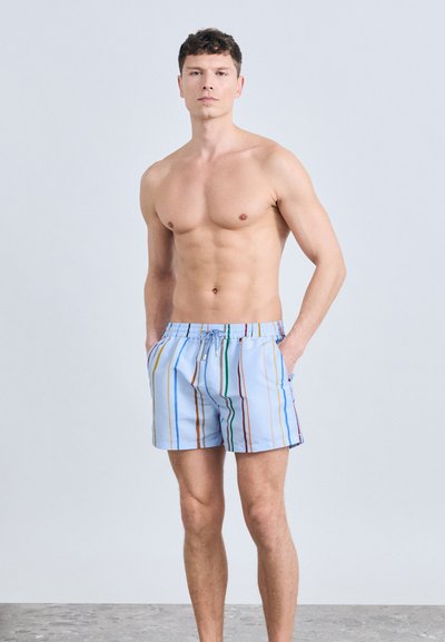 Άνδρας που στέκεται με τα χέρια στις τσέπες, φορώντας ανοιχτόχρωμα μπλε σορτς με πολύχρωμες κάθετες ρίγες σε απλό φόντο.