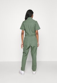 Olivengrüner Jumpsuit aus Baumwolle, mit kurzen Ärmeln, tailliertem Bund und zwei Gesäßtaschen, kombiniert mit weißen Sneakers.