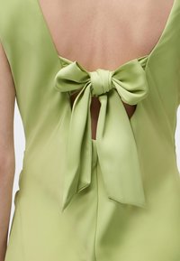 Vue arrière d'une personne portant une robe en satin vert clair avec un grand nœud noué au milieu du dos, laissant apparaître un petit espace de peau sous le nœud.