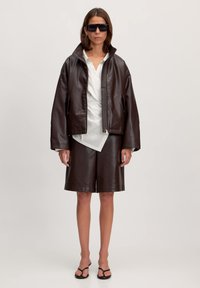 Donkerbruine leren jas met een hoge kraag, gecombineerd met bijpassende shorts. Eronder wordt een witte shirt gedragen, aangevuld met zwarte zonnebril.