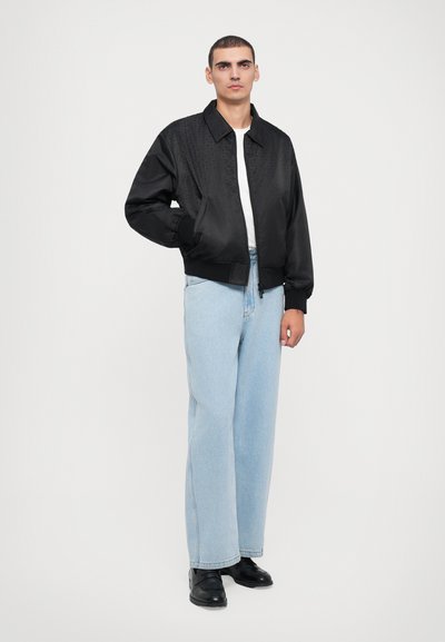 Jachetă de tip bomber black cu un model texturat în partea superioară, asociată cu blugi din denim deschis la culoare, cu croiala largă, și pantofi negri. Tricou alb simplu dedesubt.