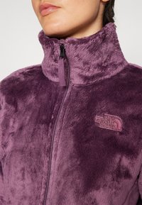 Detaljbild av en person som bär en zip-up lila fleecejacka med hög krage och The North Face-logotyp på bröstet.