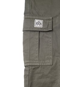 Khaki cargobyxsektion med en ficklock och en etikett med korsade element. Materialet är slitstark bomull med en mjuk yta.