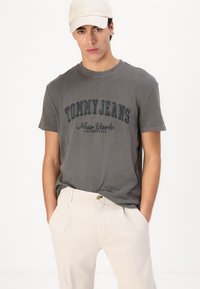 Camiseta de algodón gris con el texto "TOMMY JEANS NEW YORK" en azul oscuro, combinada con pantalones claros beige. Una gorra de béisbol neutra completa el atuendo.