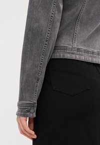 Veste en denim gris avec une coupe ajustée, des détails en boutons sur les manches et une finition texturée. Associée à un jean noir, poche légèrement visible.