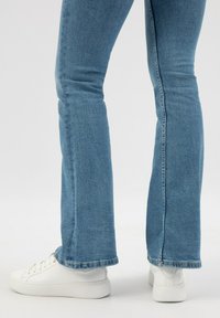 Světle modré deniminové zvonové džíny s jemným opotřebením, roztrhaným lemem a bílými teniskami s texturovanou koženou úpravou a kulatou špičkou.