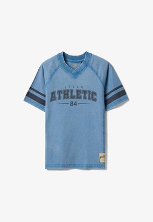 Camiseta azul de manga corta con cuello en V, con texto "Urban Athletic 84" y franjas negras en las mangas, con un pequeño parche cerca del dobladillo.
