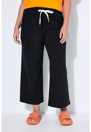 Personne portant un pantalon large noir avec un cordon blanc, un haut orange et des sandales slides orange, debout devant un fond blanc.