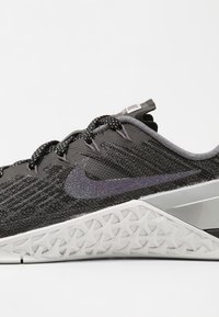 Nike lenkkari, jossa on musta verkkomateriaali yläosa, kimaltava violetti swoosh, harmaita yksityiskohtia ja muotoiltu valkoinen pohja, jossa on teksturoitu kuvio.