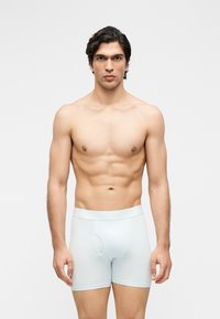 Νέος άνδρας με σκουρόχρωμα μαλλιά όρθιος, φοράει λευκά μπόξερ, σε απλό ανοιχτό γκρι φόντο.