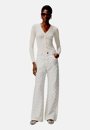Donna con cardigan crema a coste bottonato, pantaloni bianchi a gamba larga con piccole borchie, occhiali da sole, orecchini a cerchio e scarpe basse con fantasia.