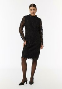 Schwarzes Spitzenkleid mit langen Ärmeln, das ein Blattmuster aufweist. Der Saum ist gewellt, und es wird mit schwarzen spitzen Pumps kombiniert.