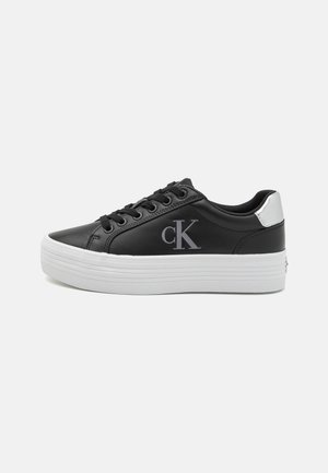 Baskets basses noires avec semelle plateforme épaisse blanche, lacets noirs, patch argenté au talon et logo "cK" sur le côté.