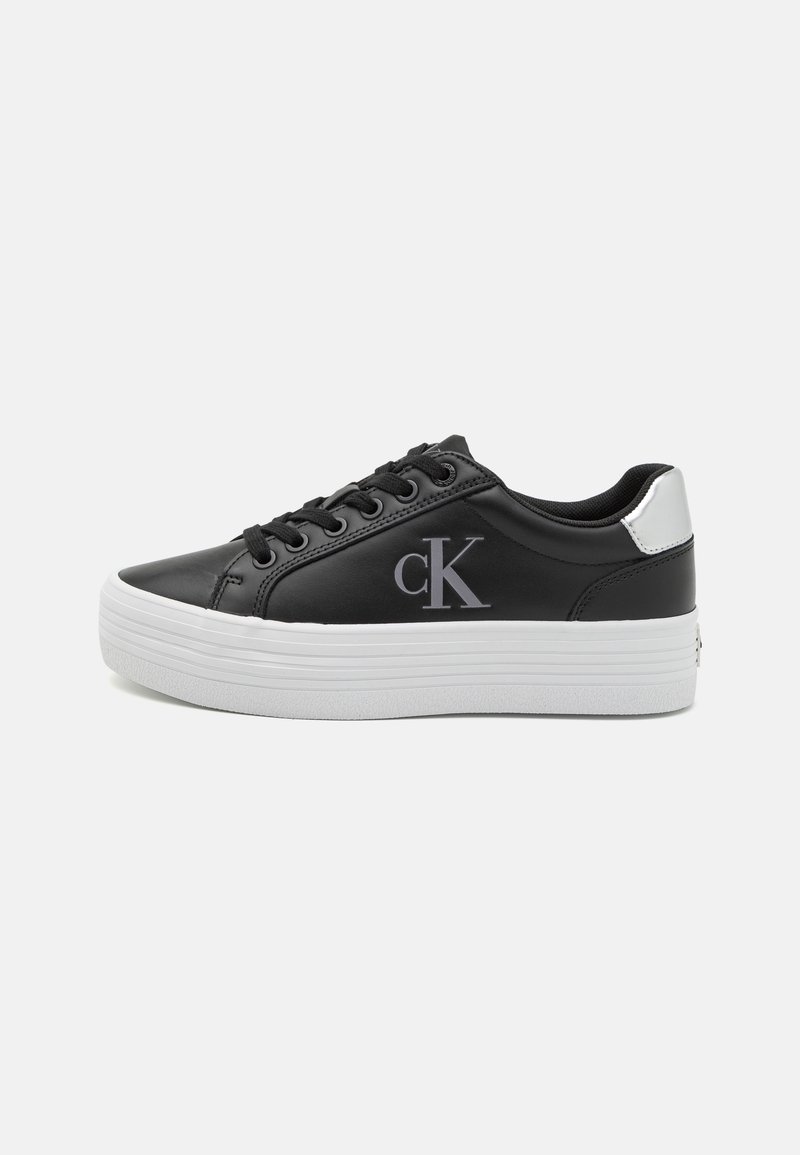 Baskets basses noires avec semelle plateforme épaisse blanche, lacets noirs, patch argenté au talon et logo "cK" sur le côté.