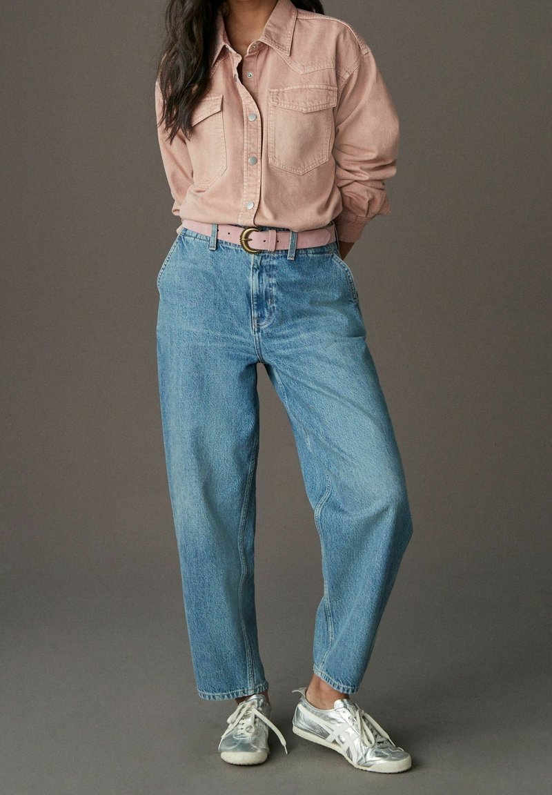 Femme portant une chemise rose clair à boutons, un jean bleu clair avec une ceinture rose et des baskets argentées, debout devant un fond uni.