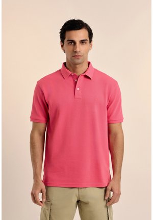 Hombre con una camisa polo de manga corta rosa y pantalones cortos cargo beige, de pie frente a un fondo claro y liso.