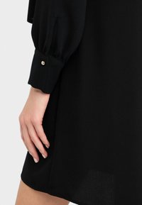 Abito nero con una lunga manica a sbuffo e polsini con bottoni. Tessuto liscio con una consistenza morbida, design minimalista e senza motivi visibili.