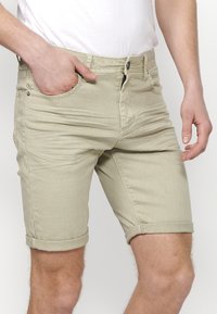 Shorts de mezclilla beige claro con dobladillos enrollados, diseño de cinco bolsillos y pasadores para cinturón. Presenta una textura sutil y cierre de botón metálico.