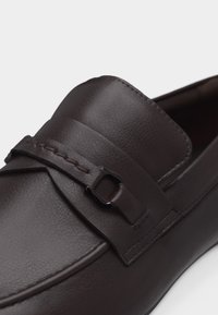 Mocassin en cuir marron foncé avec une bride cousue et un détail de boucle en métal sur la partie supérieure de la chaussure.