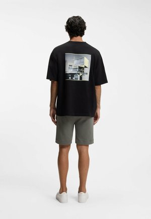 Mand med ryggen til iført sort oversized t-shirt med abstrakt moderne bygningsprint, grå shorts og hvide sneakers på hvid baggrund.