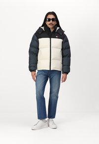 Chaqueta acolchada con colores negro y crema, alto cuello, cierre con cremallera y parche con logotipo. Combinada con jeans azules y zapatillas blancas.