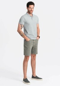 Šedé polo tričko s límečkem, olivové kraťasy a tmavě šedé nazouvací boty. Model stojí s rukama v kapsách a vystavuje tetování na pažích.