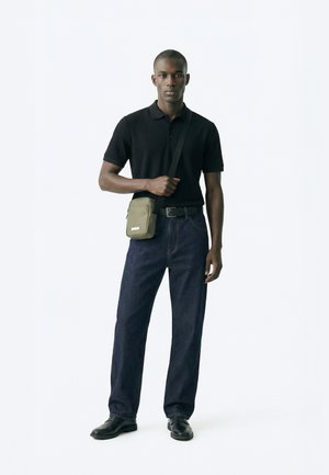 Mężczyzna ubrany w czarną koszulkę polo z krótkim rękawem, ciemnoniebieskie dżinsy, czarne buty i zieloną torbę na ramię z regulowanym paskiem na białym tle.