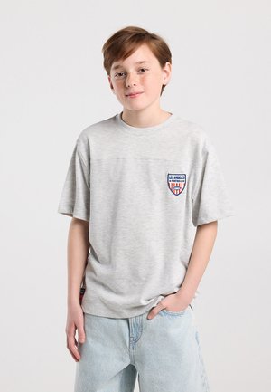 Jonge persoon met lichtbruin haar en sproeten draagt een lichtgrijs oversized T-shirt met een Los Angeles Football-patch en lichtblauwe spijkerbroek.