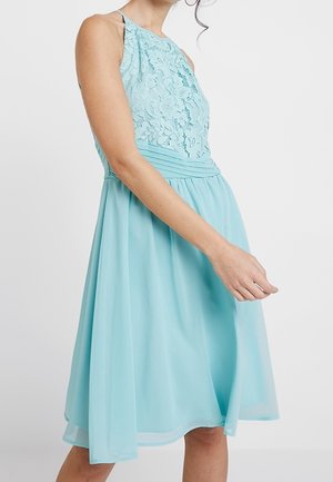 Woman wearing sleeveless light blue dress with lace bodice and pleated waist, flowing skirt falling above knees, white background.