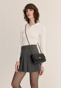 Černá kožená crossbody taška se zlatým zapínáním, doplněná vroubkovaným bílým tričkem a šedou plisovanou sukní, což podtrhuje elegantní design.